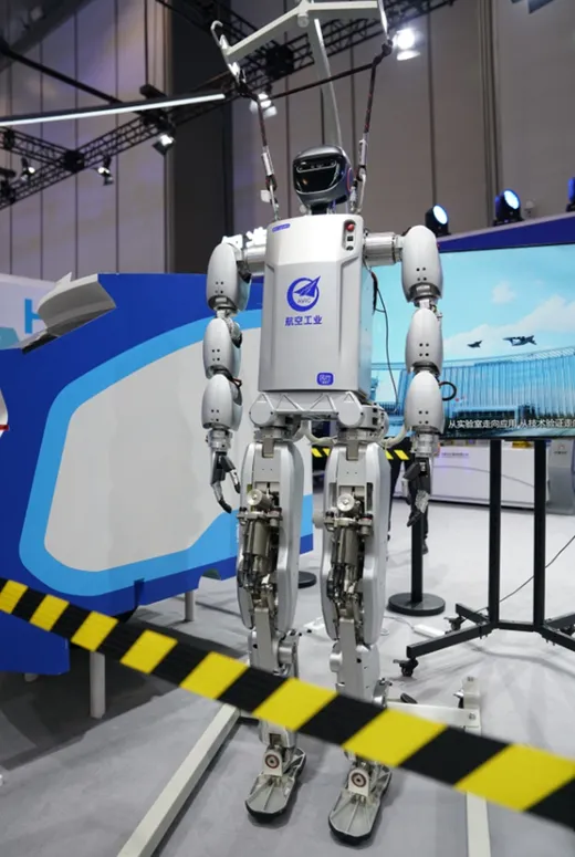 A white and silver bipedal humanoid robot with an AVIC logo on its chest is suspended by an overhead rigging harness at a WRC 2026 trade-show booth, aviation-themed graphics visible on a screen behind it, yellow-and-black caution tape in the foreground.