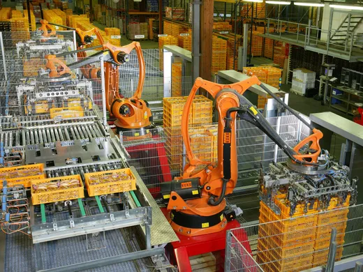 Two orange KUKA industrial robot arms palletizing bread and toast crates onto a conveyor line in a bakery warehouse