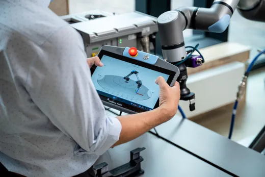 A technician in a light-blue shirt holds a Universal Robots SP7 Smart Panel displaying the PolyScope X interface with a 3D robot-arm visualization, next to a Universal Robots collaborative robot arm in a workshop setting.