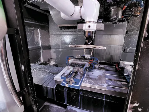 A white OB7 collaborative robot arm reaching into an active CNC milling machine coated in coolant spray, tending a vise-held workpiece