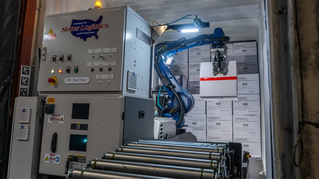 A KUKA robotic arm unloading boxed cases from inside a semi-trailer next to a States Logistics control panel, with a roller conveyor in the foreground.