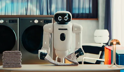 A white wheeled robot with articulated arms and a rounded head displaying two 'C' shaped digital eyes, an LG logo on its torso, folds a towel on a countertop next to two front-loading washing machines and a stack of folded towels in a home laundry room.