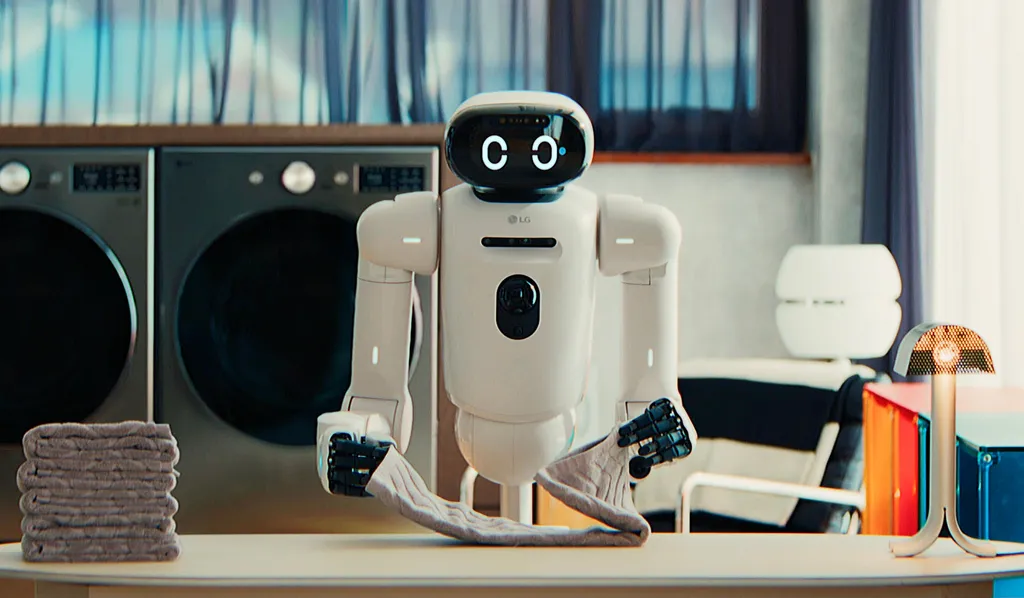 A white wheeled robot with articulated arms and a rounded head displaying two 'C' shaped digital eyes, an LG logo on its torso, folds a towel on a countertop next to two front-loading washing machines and a stack of folded towels in a home laundry room.