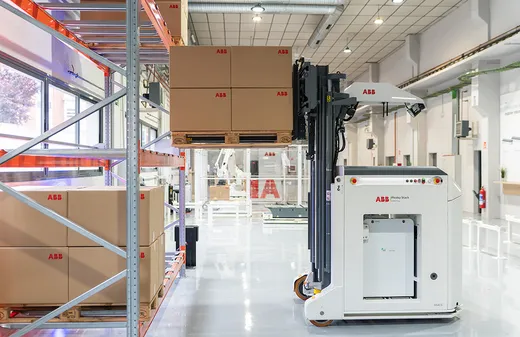 A white ABB Flexley Stack F712 autonomous forklift raises its mast to place a pallet of ABB-branded cardboard boxes onto steel warehouse racking, with more loaded racking to the left and robotics cells visible in the background.