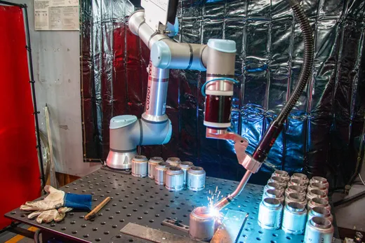 A Universal Robots-based cobot arm labeled 'Cobot Welder' performs MIG welding on small cylindrical metal parts on a perforated welding table, sparks flying, with welding gloves and a wire brush resting nearby and a metallic welding curtain in the background.