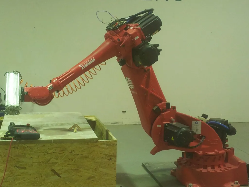 A red six-axis Comau industrial robot arm with its gripper extended over a wooden platform in a workshop