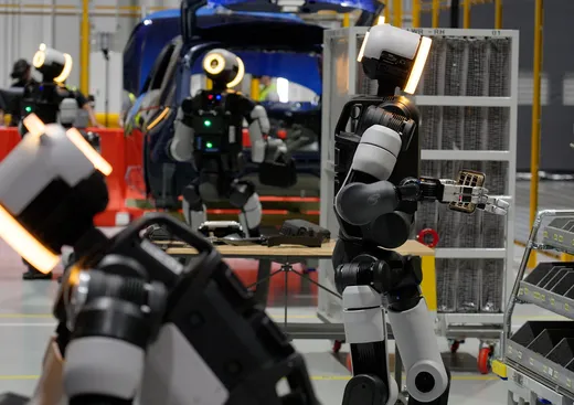 Several humanoid Atlas robots with glowing amber head lights work at tables and parts racks beside a blue vehicle body on a factory floor.