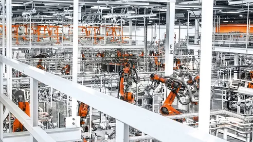 Orange KUKA industrial robot arms positioned along an elevated automotive body-in-white production line, with a catwalk and metal railing visible in the foreground.