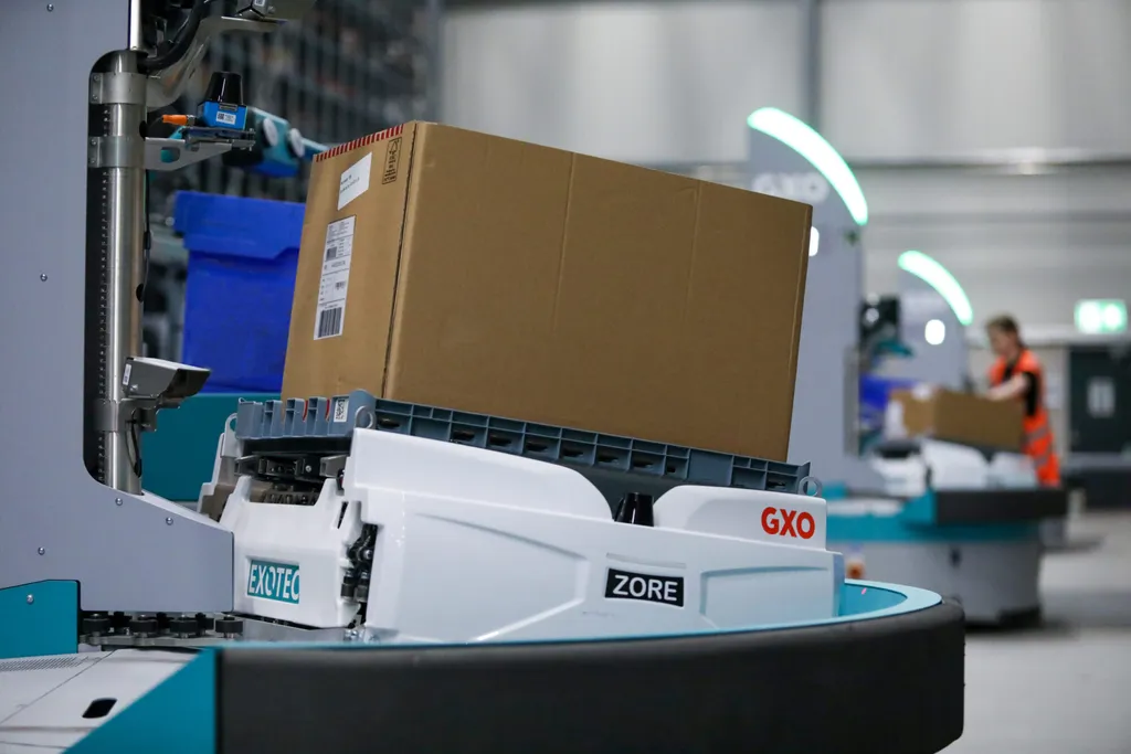Close-up of a teal-and-white Exotec 'Zore' goods-to-person robot branded EXOTEC and GXO lifting a cardboard shipping box onto a conveyor turntable, with a warehouse worker in an orange vest and additional Skypod robot towers with green status-light arcs visible in the blurred background.