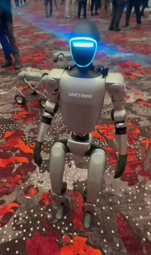 Unitree's G1 humanoid robot, with UNITREE branding and a glowing blue face display, standing on a trade-show expo floor.