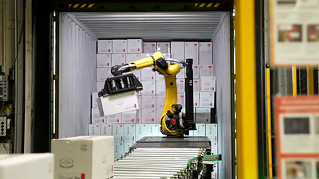A yellow robotic arm reaching into a shipping container full of cartons to unload a boxed case onto a roller conveyor.