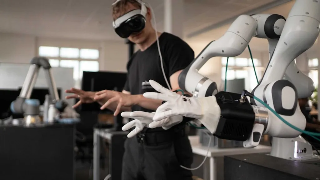 A man wearing a VR headset and white glove-based teleoperation controllers, reaching toward a white-and-black six-axis robotic arm fitted with a dexterous gripper, in an office/lab setting.