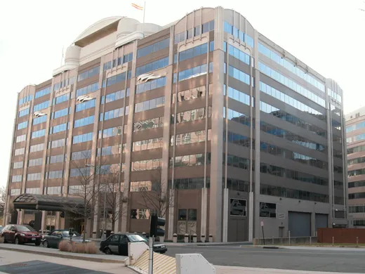 Exterior street-level view of the FCC headquarters building in Washington, D.C. — an art-deco-influenced modern office building with cars parked out front and bare winter trees.