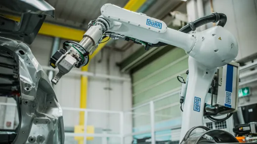 A white robotic arm branded DÜRR in blue lettering, working on a silver automotive body-in-white inside a factory bay with yellow safety railing and a green roll-up door in the background.