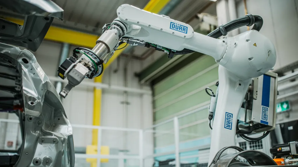 A white robotic arm branded DÜRR in blue lettering, working on a silver automotive body-in-white inside a factory bay with yellow safety railing and a green roll-up door in the background.