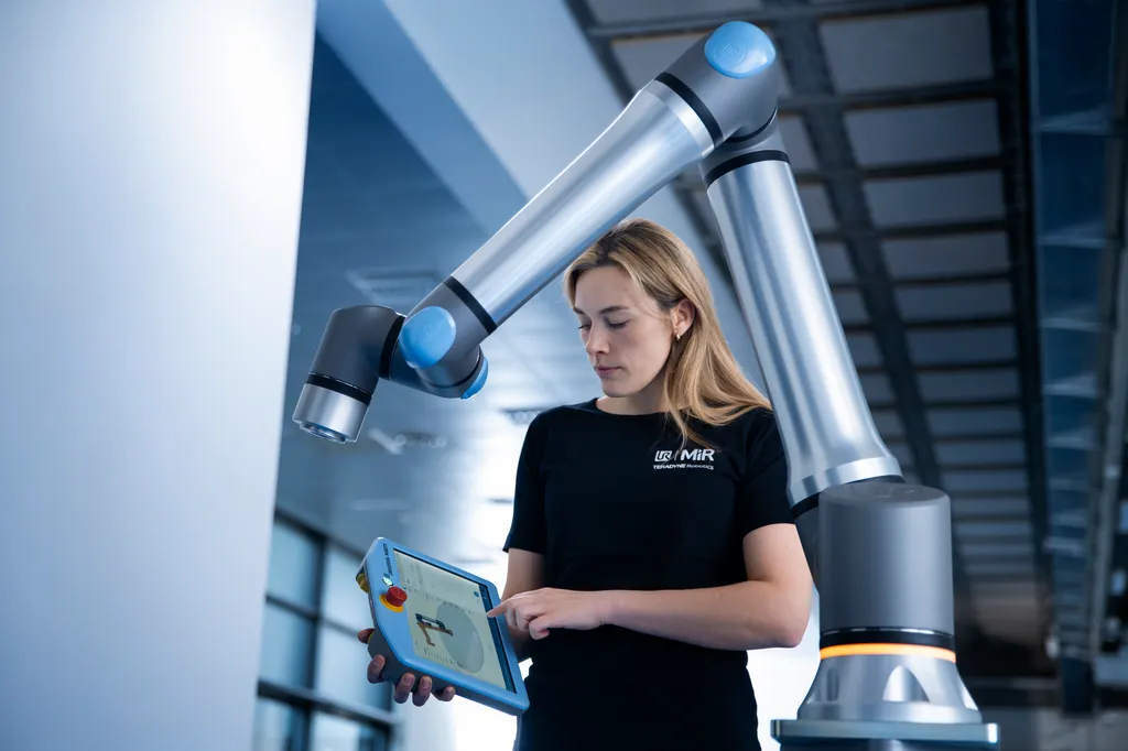 A technician in a UR / MiR / Teradyne Robotics branded shirt stands beneath a raised Universal Robots collaborative arm, programming it with a handheld blue teach pendant.