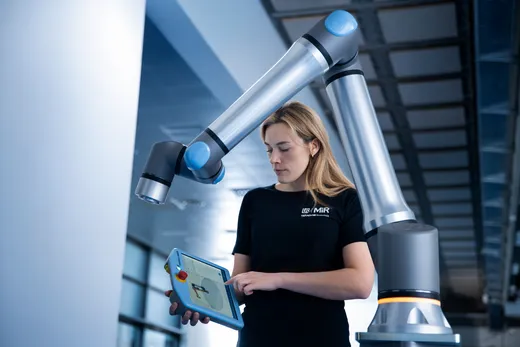 A technician in a UR / MiR / Teradyne Robotics branded shirt stands beneath a raised Universal Robots collaborative arm, programming it with a handheld blue teach pendant.
