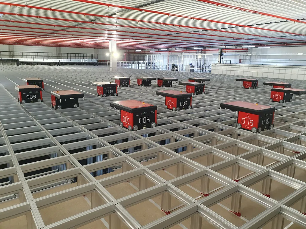 Numbered red-and-black AutoStore robots sit atop an aluminum storage grid in a warehouse, with rows of storage bins visible beneath the grid cells.