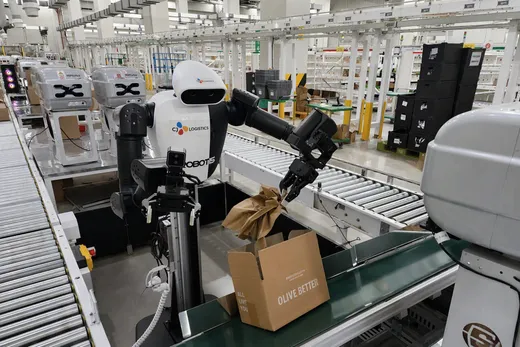 A stationary dual-arm humanoid robot branded CJ Logistics and Robotis uses one gripper to insert crumpled brown cushioning paper into an Olive Young-branded shipping box on a roller conveyor packaging line, with automated parcel sorters visible in the background.