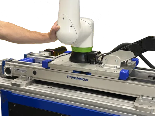 A technician's hand positions a white and green FANUC CRX collaborative robot arm mounted on a blue-and-silver Thomson Movotrak linear rail unit, with THOMSON branding visible on the rail.