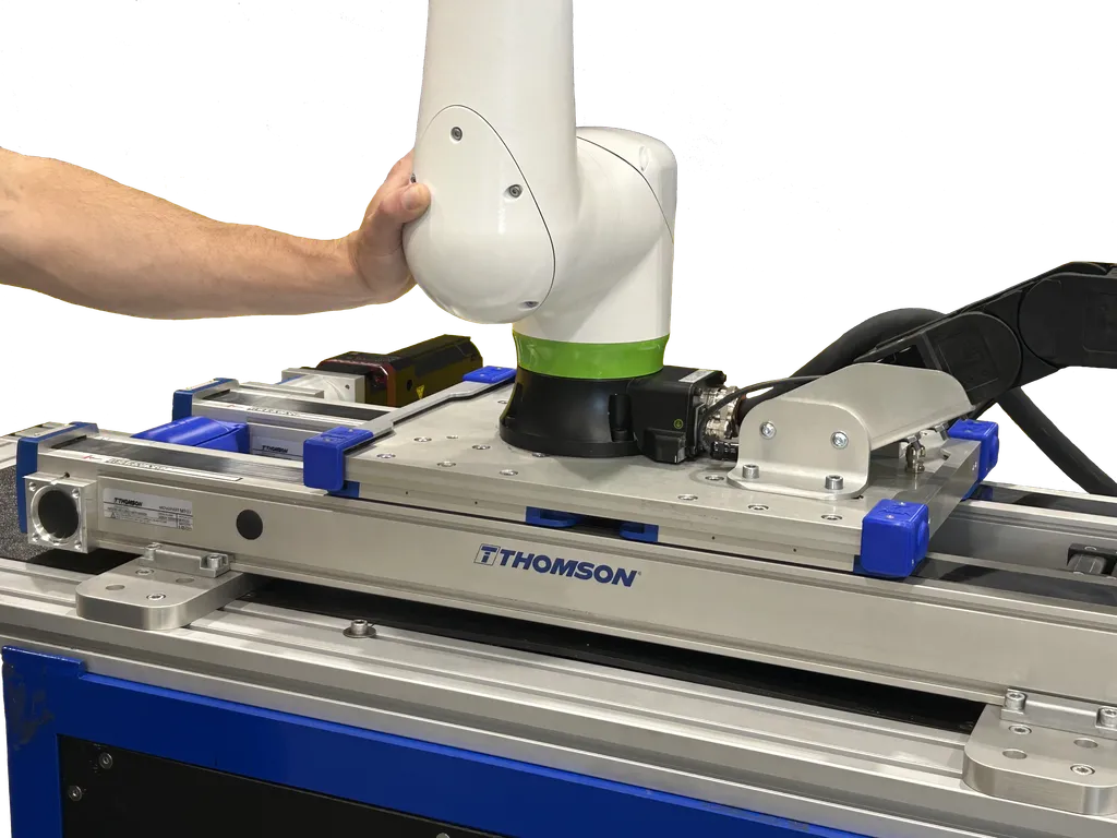 A technician's hand positions a white and green FANUC CRX collaborative robot arm mounted on a blue-and-silver Thomson Movotrak linear rail unit, with THOMSON branding visible on the rail.