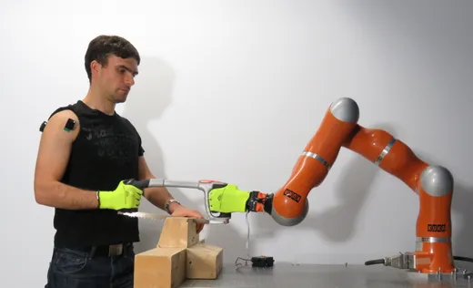 A researcher uses a handsaw jointly gripped with an orange KUKA collaborative robot arm during a human-robot-collaboration lab demonstration.