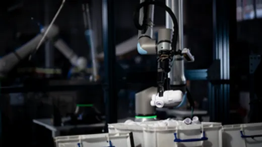 A robotic gripper arm reaching down into small parts bins on an industrial automation cell, with a second robotic arm visible in the dimly lit background