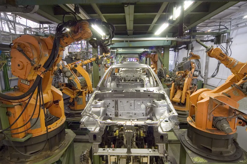 Orange KUKA industrial robot arms on both sides of an unfinished car body shell on an automotive welding line