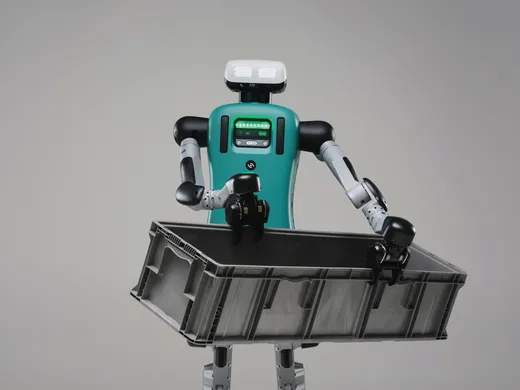 Agility Robotics' Digit humanoid — a bipedal robot with a teal torso, a white LED face panel and articulated black-and-silver arms — carries a large gray warehouse tote against a plain studio backdrop.