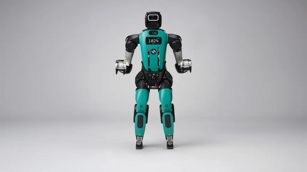 A teal-and-black bipedal humanoid robot with human-like legs stands facing forward against a plain gray studio backdrop, its chest display reading '1024 AUTO' and three-fingered grippers hanging at its sides.