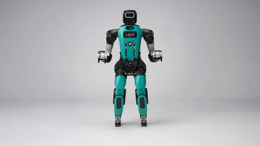A teal-and-black bipedal humanoid robot with human-like legs stands facing forward against a plain gray studio backdrop, its chest display reading '1024 AUTO' and three-fingered grippers hanging at its sides.
