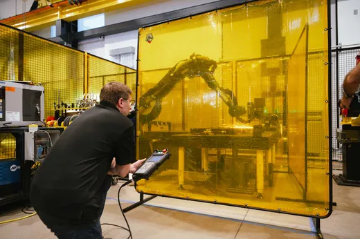 A technician in a black polo shirt holds a robot teach pendant while operating a yellow-branded Yaskawa six-axis robotic arm performing arc welding inside a yellow-curtained safety cell, with a Miller welding power supply visible in the foreground.