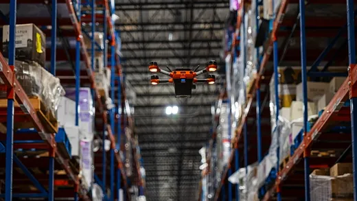 An orange-and-black Corvus One quadcopter drone hovers at height in a dim warehouse aisle between tall pallet racking loaded with shrink-wrapped cartons, with skylights and ceiling lighting visible above.